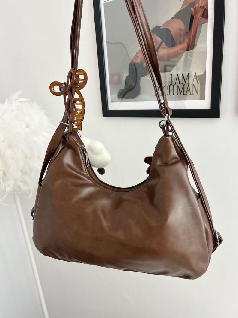 May include: A brown hobo-style shoulder bag with a smooth finish, featuring a top zipper closure and long adjustable straps. A translucent amber-colored claw clip is attached to one of the shoulder straps, adding a decorative element to the bag.