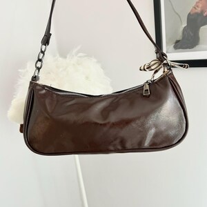 May include: A brown baguette-style shoulder bag with a zipper closure and a thin strap. The bag has a shiny, smooth texture and a small decorative scarf tied to the strap. A chain detail is visible on the strap. The bag is hanging in front of a white wall.