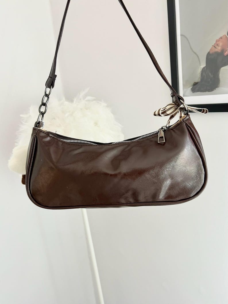 May include: A brown baguette-style shoulder bag with a zipper closure and a thin strap. The bag has a shiny, smooth texture and a small decorative scarf tied to the strap. A chain detail is visible on the strap. The bag is hanging in front of a white wall.