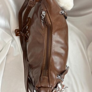 May include: A brown backpack with a smooth finish, featuring a vertical zippered main compartment, silver-toned metal hardware, adjustable shoulder straps, and a small decorative charm attached to the side zipper pull near the base.