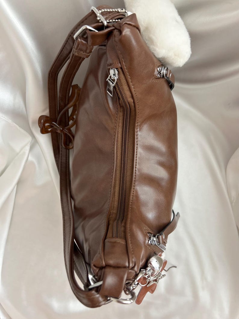 May include: A brown backpack with a smooth finish, featuring a vertical zippered main compartment, silver-toned metal hardware, adjustable shoulder straps, and a small decorative charm attached to the side zipper pull near the base.