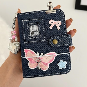 Può includere: Una copertina per taccuino in denim blu scuro con una clip in metallo argentato sulla parte superiore, una piccola finestra per foto, una toppa a forma di fiocco rosa, una grande toppa a farfalla rosa con ciondolo a cuore, una stella e un portachiavi.