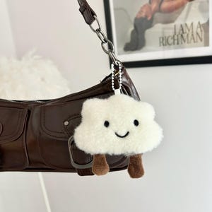 May include: A brown leather shoulder bag with a silver chain strap. A white fluffy cloud-shaped keychain with a smiling face and brown legs hangs from the bag. The background includes a framed print with the text "I AM A RICH MAN".