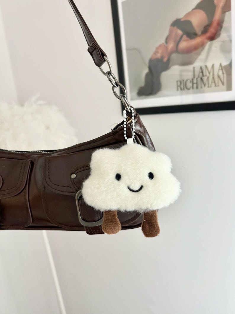 May include: A brown leather shoulder bag with a silver chain strap. A white fluffy cloud-shaped keychain with a smiling face and brown legs hangs from the bag. The background includes a framed print with the text "I AM A RICH MAN".