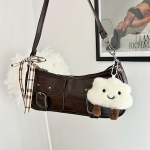 May include: A brown leather shoulder bag with a long strap and a small front pocket. The bag features a silver buckle and a white cloud-shaped keychain with brown legs. A brown and white checkered ribbon is tied to the strap.