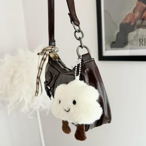 May include: A brown leather shoulder bag with a matching strap and silver-tone hardware. A fluffy white cloud-shaped keychain with brown feet hangs from the bag. The bag has a curved shape and a small bow detail.