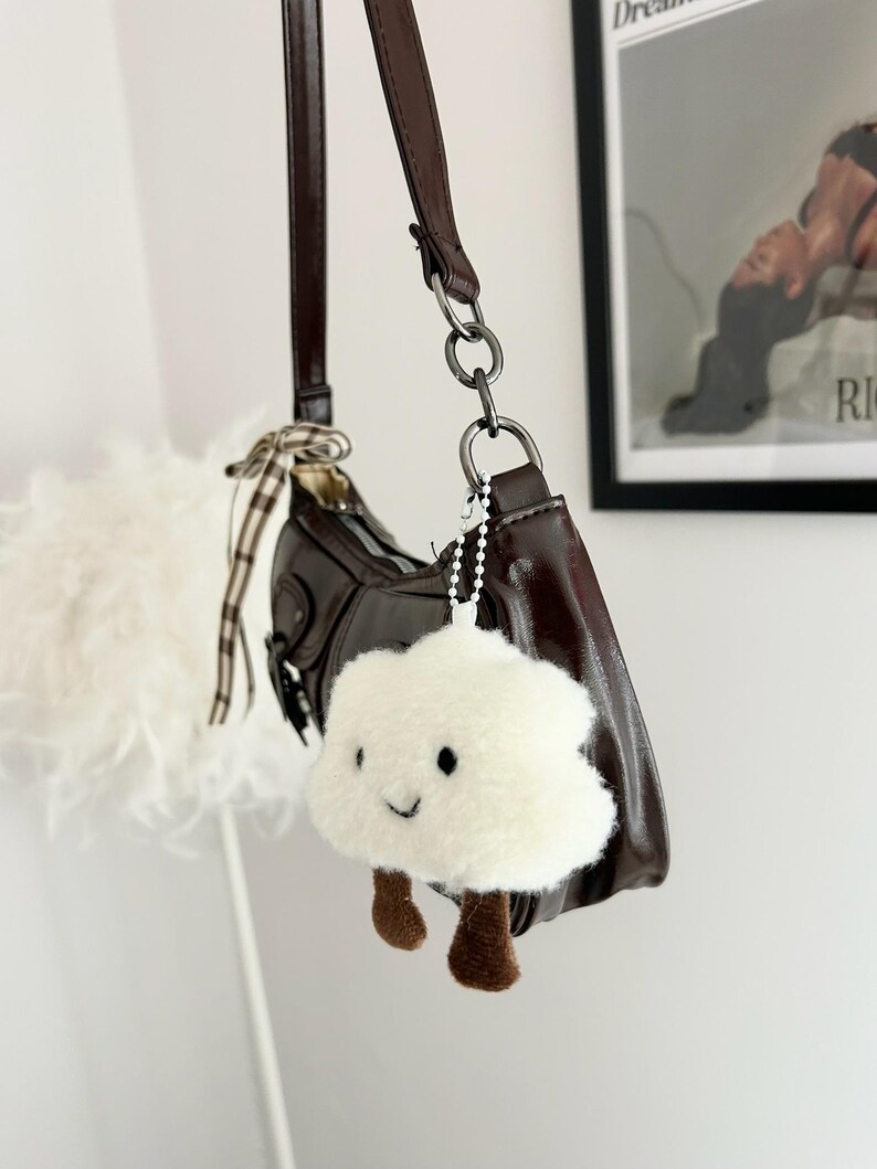 May include: A brown leather shoulder bag with a matching strap and silver-tone hardware. A fluffy white cloud-shaped keychain with brown feet hangs from the bag. The bag has a curved shape and a small bow detail.
