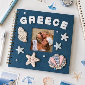 May include: A square, spiral-bound photo album with a dark blue fabric cover featuring the word GREECE in white letters. The cover is decorated with beaded seashell and starfish appliqués, small white pearls, and a central square photo window.