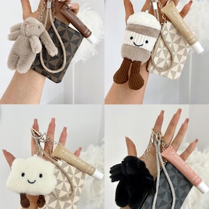 May include: Four keychains with small plush figures, mini wallets, and lip butter balm tubes. The keychains feature a bunny, a coffee cup, a cloud, and a black fluffy figure. The wallets have patterned designs, and the lip balm tubes are beige.