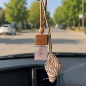 May include: A car air freshener hanging from a tan cord, featuring a square glass bottle with a wooden cap and a label that reads SHELL, YEAH. Next to the bottle hangs a decorative shell-shaped charm covered in small white beads and gold trim.