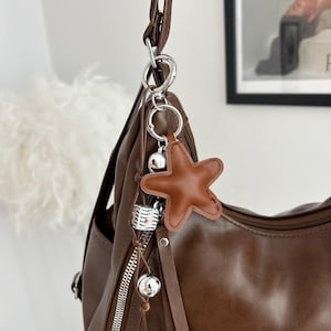 May include: A brown shoulder bag featuring a silver metal zipper and a decorative keychain. The keychain includes a brown star-shaped charm, a small white and black patterned cube, and two silver spherical beads hanging from a thin brown cord.