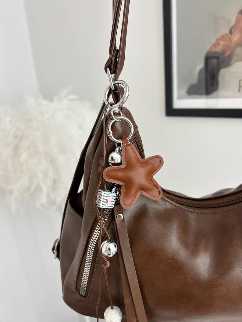 May include: A brown shoulder bag featuring a silver metal zipper and a decorative keychain. The keychain includes a brown star-shaped charm, a small white and black patterned cube, and two silver spherical beads hanging from a thin brown cord.