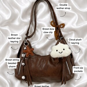 May include: A brown leather shoulder bag featuring double leather straps, front zip pockets with brown leather tassel pulls, and a brown string beaded charm. Accessories include a brown leather star keyring, a cloud plush keyring, and a brown bow claw clip.