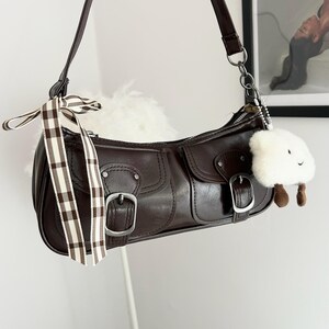 May include: A brown leather shoulder bag with silver hardware, two front pockets, and buckle closures. A decorative ribbon and a white cloud-shaped charm hang from the bag. The bag has a long, adjustable strap.