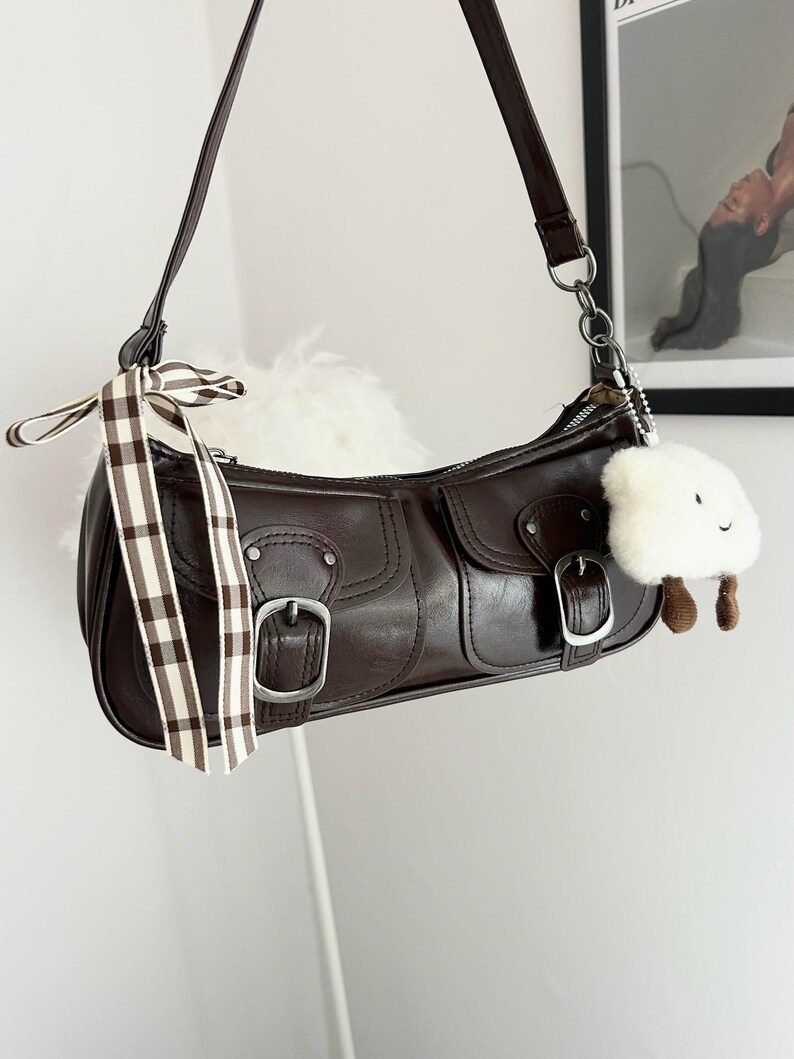 May include: A brown leather shoulder bag with silver hardware, two front pockets, and buckle closures. A decorative ribbon and a white cloud-shaped charm hang from the bag. The bag has a long, adjustable strap.