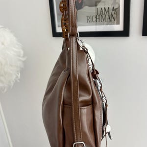 May include: A brown shoulder bag with a smooth finish, featuring a long adjustable strap with a silver-toned metal buckle, a side pocket, and a decorative charm attachment with silver-toned hardware and small white spherical accents hanging from the side.