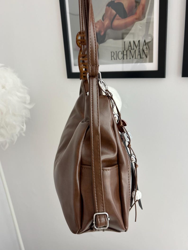 May include: A brown shoulder bag with a smooth finish, featuring a long adjustable strap with a silver-toned metal buckle, a side pocket, and a decorative charm attachment with silver-toned hardware and small white spherical accents hanging from the side.