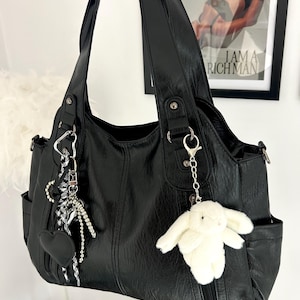 May include: Black faux leather tote bag with multiple compartments and silver-tone hardware. The bag has a white plush bunny keyring and a black heart charm. A framed print with the text "I AM A RICH MAN" is visible in the background.