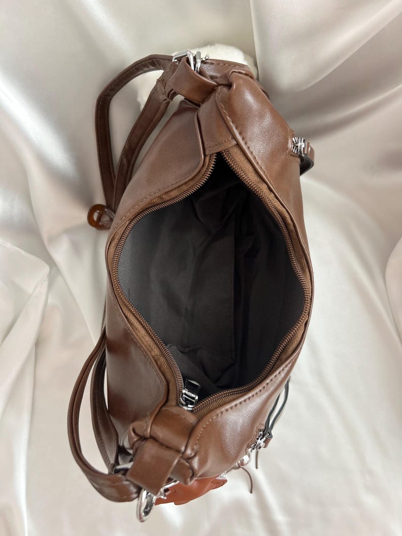 May include: A brown shoulder bag with a smooth finish, featuring a top zipper closure that opens to reveal a dark gray interior lining. The bag includes silver-toned metal hardware, a matching brown strap, and additional side zipper details.