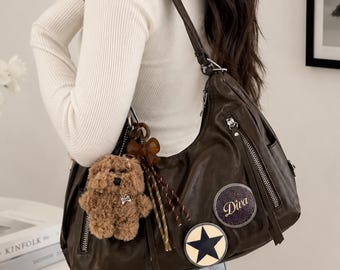 May include: A dark brown hobo-style shoulder bag featuring silver-tone zippers, a plush brown teddy bear keychain, and two circular patches. One patch displays a black star on a cream background, while the other features the word Diva in script.