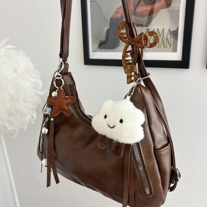 May include: A brown shoulder bag with a smooth finish, featuring a long strap, silver-tone metal zippers, and decorative hanging charms. The charms include a brown star, a white plush cloud with a smiling face, and a brown tortoiseshell hair clip.