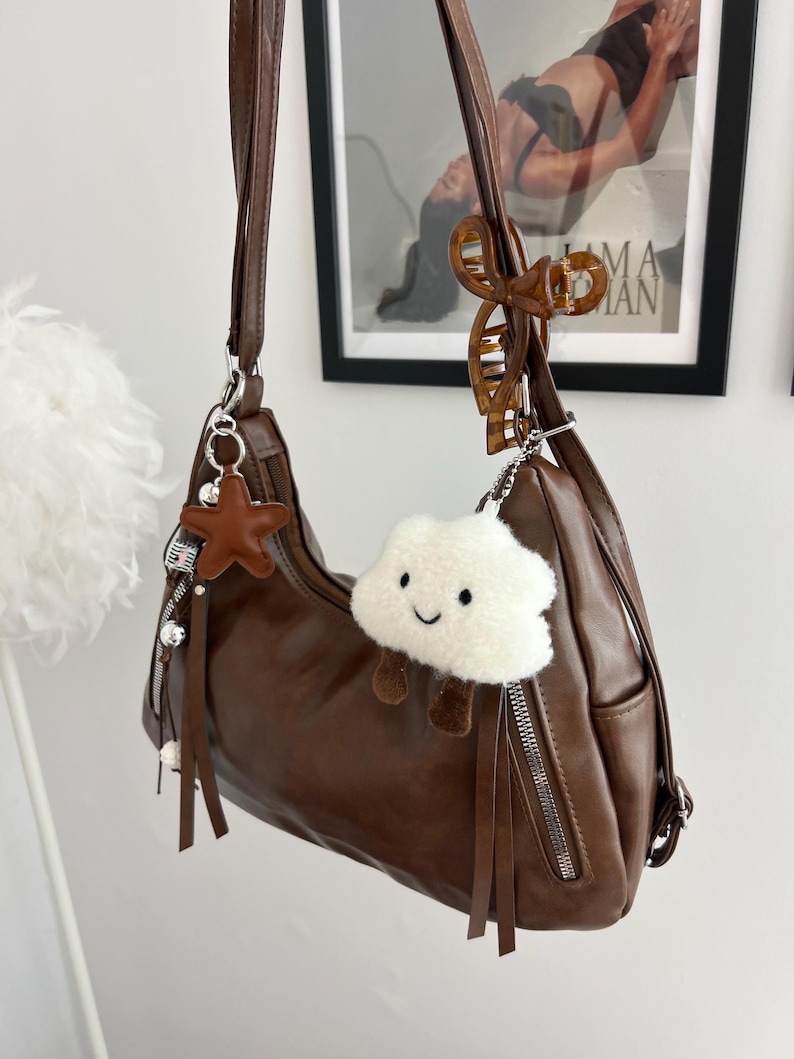 May include: A brown shoulder bag with a smooth finish, featuring a long strap, silver-tone metal zippers, and decorative hanging charms. The charms include a brown star, a white plush cloud with a smiling face, and a brown tortoiseshell hair clip.