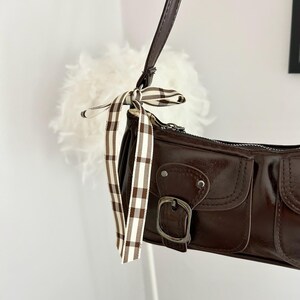 May include: A brown shoulder bag with a glossy finish. The bag features two front pockets with silver buckles and a decorative ribbon with a brown and white plaid pattern. The strap is a dark brown color.