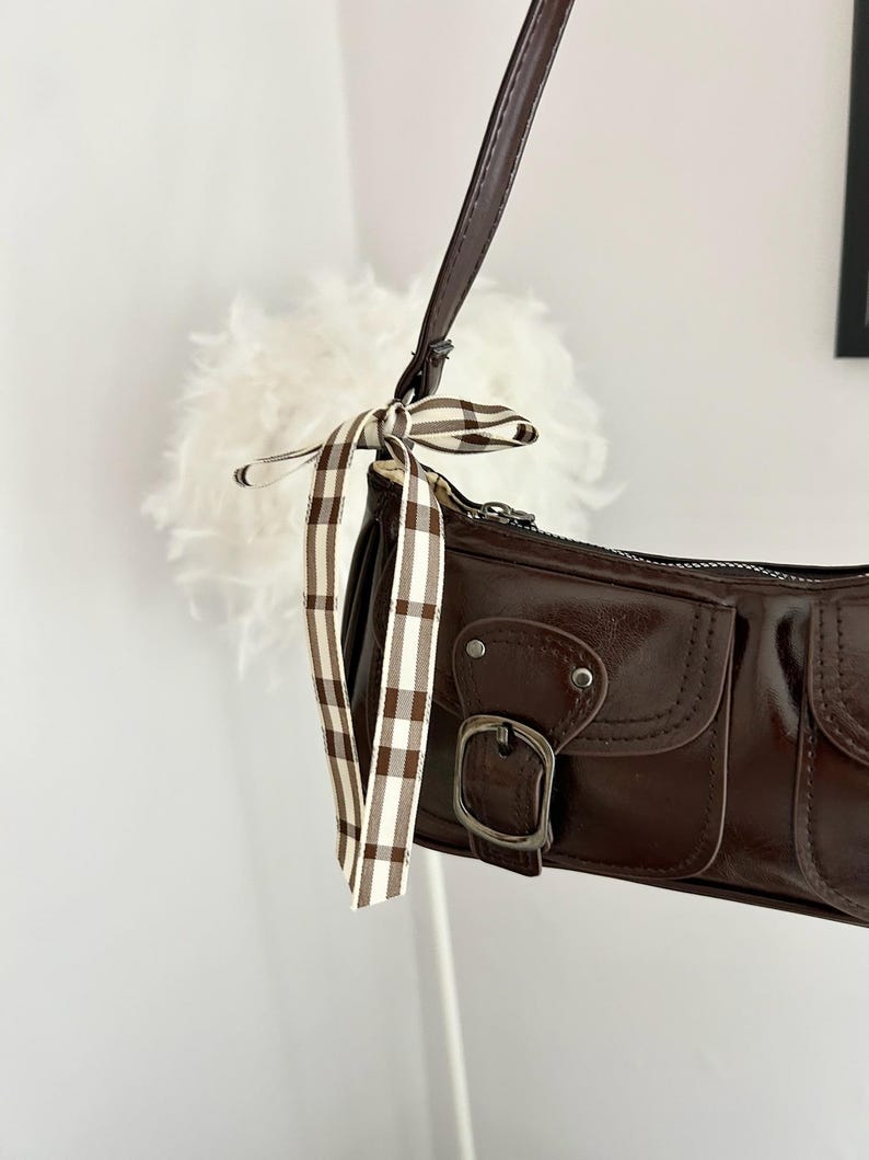 May include: A brown shoulder bag with a glossy finish. The bag features two front pockets with silver buckles and a decorative ribbon with a brown and white plaid pattern. The strap is a dark brown color.