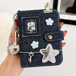 Pode incluir: Uma carteira de ganga azul-escura com uma janela retangular para fotografias, dois emblemas bordados de estrelas azul-claras e uma estrela grande de lantejoulas. Inclui um clipe de metal, um fecho de mola e um pendente de coração e chave.