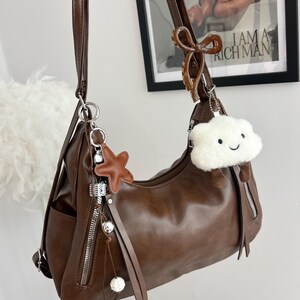 May include: A dark brown shoulder bag with a smooth finish, featuring a long adjustable strap, two vertical zippered front pockets, and decorative charms including a brown star, a white fuzzy cloud with a face, and a brown bow clip.