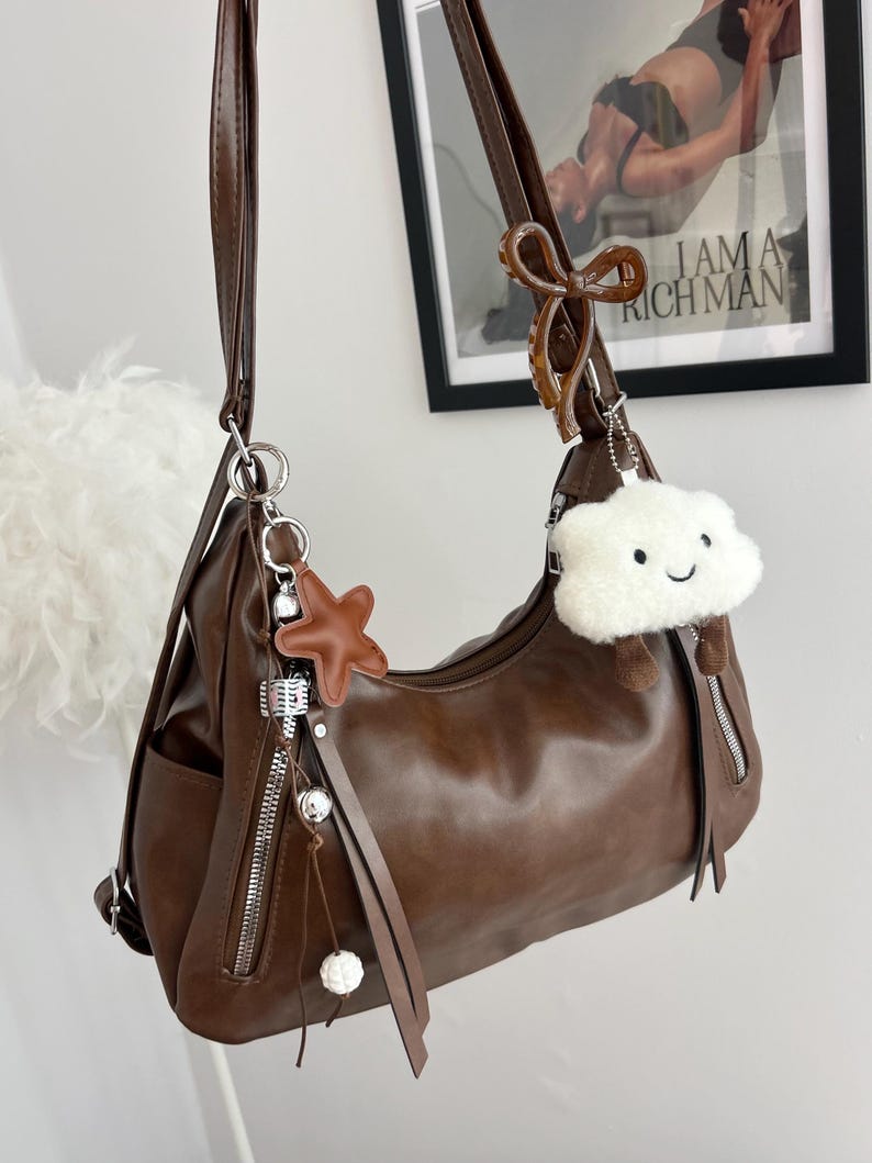 May include: A dark brown shoulder bag with a smooth finish, featuring a long adjustable strap, two vertical zippered front pockets, and decorative charms including a brown star, a white fuzzy cloud with a face, and a brown bow clip.