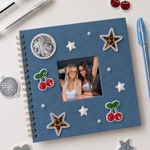 May include: A square blue denim-textured photo album with a spiral binding, featuring a central window displaying a photograph of two people. The cover is decorated with sequined cherry patches, leopard print star patches, and small white pearls.