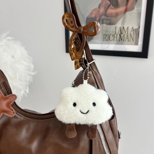 May include: A brown leather shoulder bag featuring a brown tortoiseshell bow hair clip attached to the strap and a white fuzzy cloud-shaped keychain with a smiling face and dangling brown legs hanging from a silver ball chain.