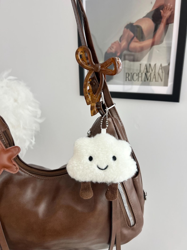 May include: A brown leather shoulder bag featuring a brown tortoiseshell bow hair clip attached to the strap and a white fuzzy cloud-shaped keychain with a smiling face and dangling brown legs hanging from a silver ball chain.