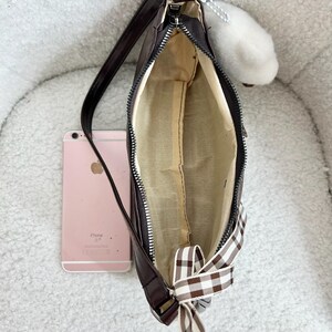 May include: A brown shoulder bag with a zipper closure and a gingham bow. The bag has a long strap and a light-colored interior. A pink smartphone is next to the bag. A white fluffy charm hangs from the zipper.