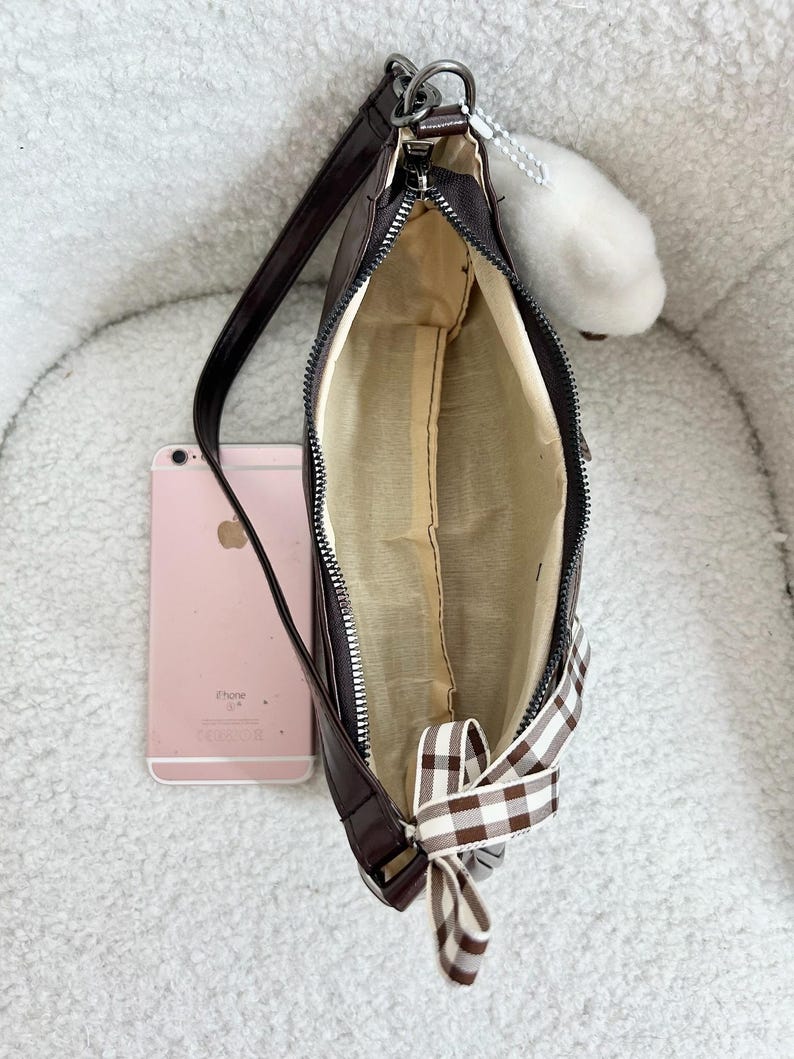 May include: A brown shoulder bag with a zipper closure and a gingham bow. The bag has a long strap and a light-colored interior. A pink smartphone is next to the bag. A white fluffy charm hangs from the zipper.