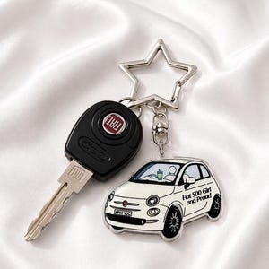 May include: A silver star-shaped keyring attached to a black Fiat car key and a white acrylic charm featuring a white Fiat 500 car illustration. The charm displays the text Fiat 500 Girl and Proud on the side and shows a driver holding a green drink.