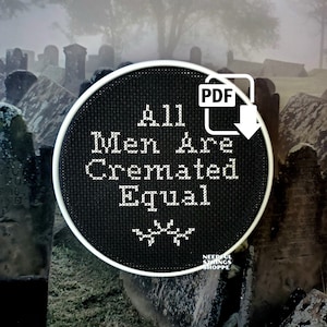 May include: A black and white cross-stitch embroidery hoop with the text "All Men Are Cremated Equal" and a skull and crossbones symbol. The hoop is in front of a blurry background of a graveyard.