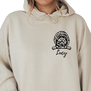May include: A beige hoodie with a black graphic of a dog peeking over the top and the name "Lucy" printed below.