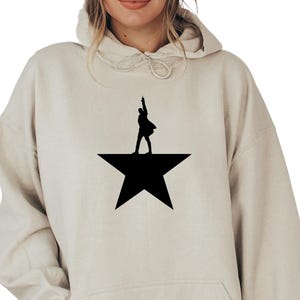 May include: A cream-colored hoodie with a black silhouette of a person standing on a star. The star is a five-pointed star.