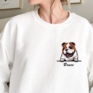 May include: White sweatshirt with a cartoon bulldog peeking over the top of a pocket. The bulldog is brown and white with a pink tongue. The text "Bruce" is printed below the bulldog.