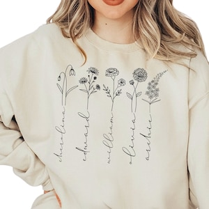 May include: A cream-colored sweatshirt featuring a floral design with names. The black line art depicts various flowers, each paired with a name: Carolina, Edward, William, Olivia, and Archie. The sweatshirt is a casual, comfortable style.