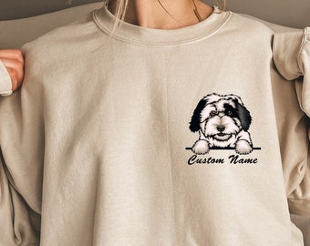 May include: A beige sweatshirt with a cartoon image of a black and white dog peeking over the edge of a box. The text "Custom Name" is printed below the image.