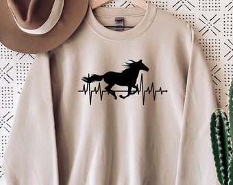 May include: A beige sweatshirt with a black silhouette of a horse running with a heartbeat graphic. The text "swipe for more colors" is visible on the right side of the image.