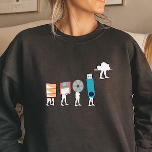 May include: Black sweatshirt with a graphic of a cloud carrying a USB drive, followed by a series of figures carrying different storage devices, including a floppy disk, a CD, and a hard drive. The text "The Evolution of Storage" is printed below the graphic.