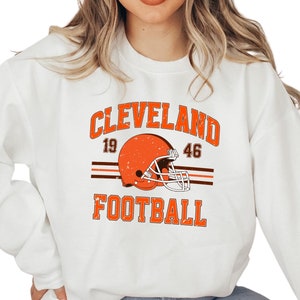 May include: White sweatshirt with a vintage-style Cleveland Browns football graphic. The graphic features a brown helmet with a white stripe, the text "CLEVELAND" in brown, and the numbers "19" and "46" in brown. The text "FOOTBALL" is in brown below the helmet.