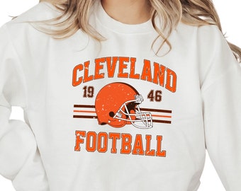 May include: White sweatshirt with a vintage-style Cleveland Browns football graphic. The graphic features a brown helmet with a white stripe, the text "CLEVELAND" in brown, and the numbers "19" and "46" in brown. The text "FOOTBALL" is in brown below the helmet.