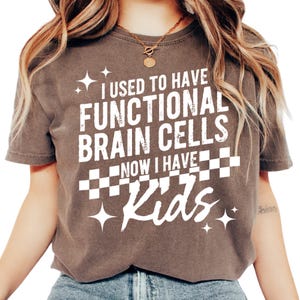 May include: A brown short-sleeve t-shirt featuring white text that reads I USED TO HAVE FUNCTIONAL BRAIN CELLS NOW I HAVE Kids. The design includes a checkered pattern and star graphics. The shirt has a crew neckline and a relaxed fit.