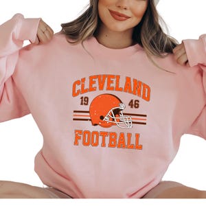 May include: A pink sweatshirt with a vintage-style graphic of a football helmet and the text "CLEVELAND FOOTBALL" with the numbers "19" and "46" on either side of the helmet.