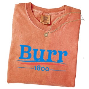May include: A coral-colored t-shirt with the word "Burr" in large blue letters, a blue line, a red line, and the number "1800" in blue. A gold necklace with a pendant rests on the shirt. The shirt has a Comfort Colors tag.